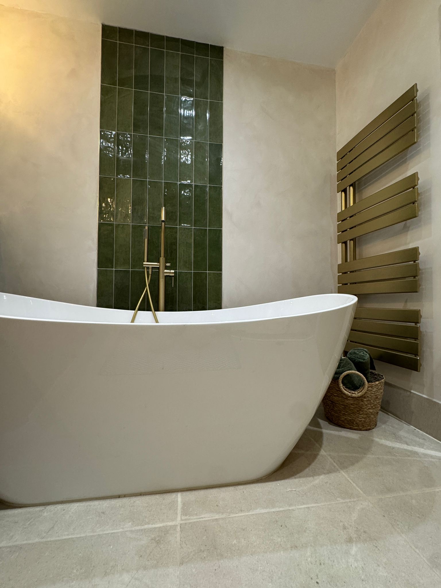 A freestanding slipper bath set against a green tiled column, with a brushed-brass floor-mounted mixer.