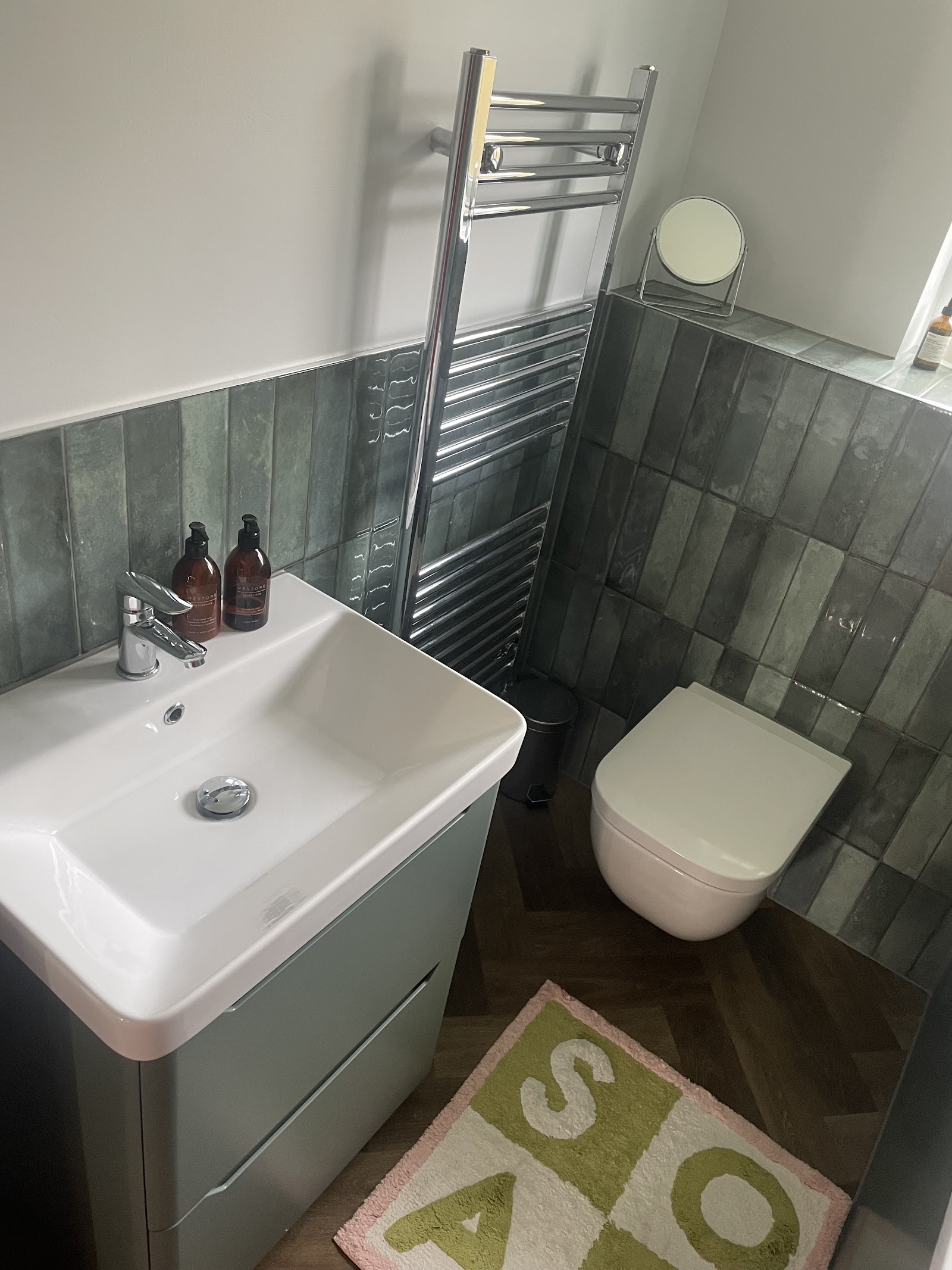 An en-suite with grey-green tiling, wall-hung WC, chrome heated towel rail and herringbone floor.